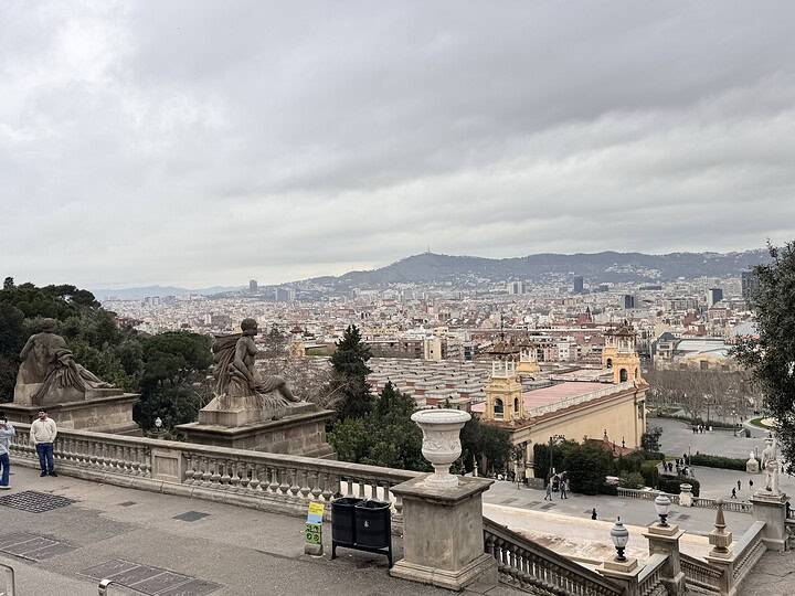 Enjoy a breathtaking view of Barcelona city from a terrace with statues and a cloudy sky, featuring yellow buildings and distant hills—the perfect start or finish to your MSC Fantasia Cruise or family 7-day itinerary.
