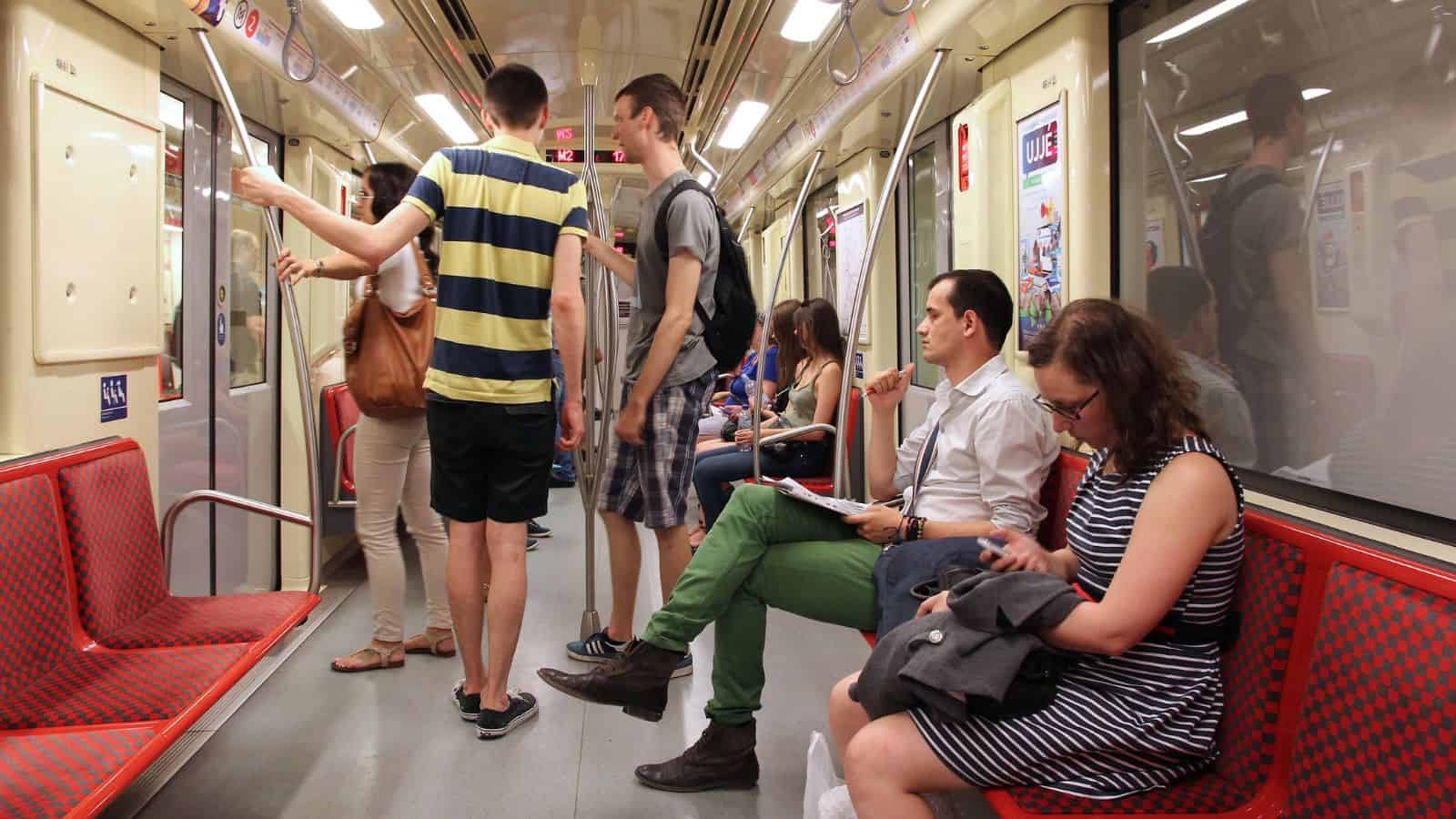 Several people are enjoying a train ride, some sitting on red-patterned seats reading or checking their phones, while others follow train etiquette by standing or preparing to exit. The well-lit interior features posters on the walls.