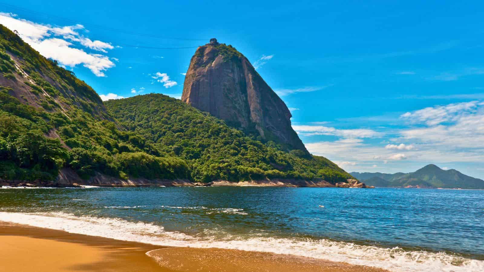 A sandy beach with ocean waves and a large green mountain under a bright blue sky.