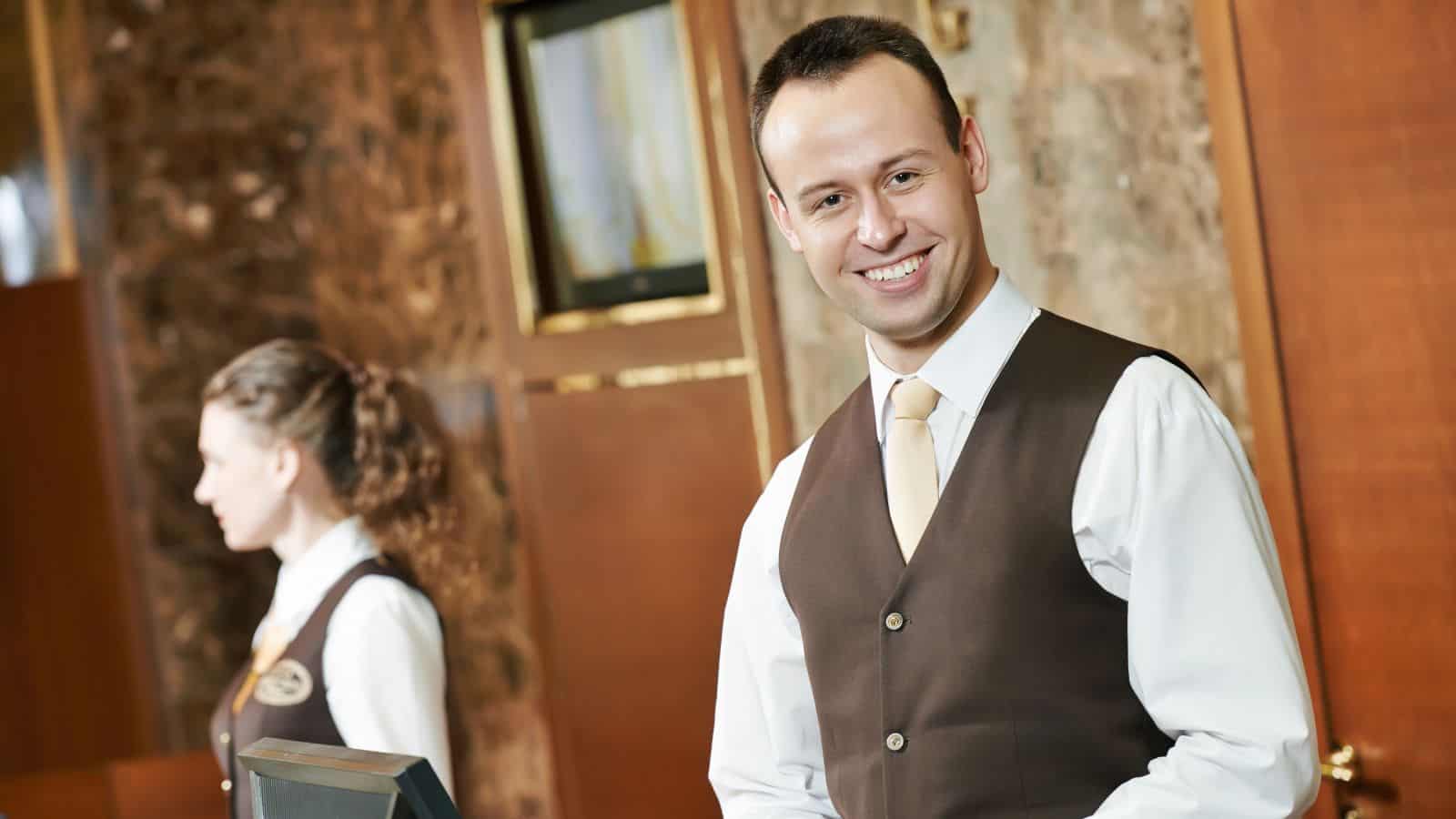 A man in a brown vest and yellow tie stands smiling at a hotel reception desk, welcoming travelers. A woman in similar attire stands in the background. The brown-walled lobby hints at comfort, perfect for those seeking all-inclusive vacations.