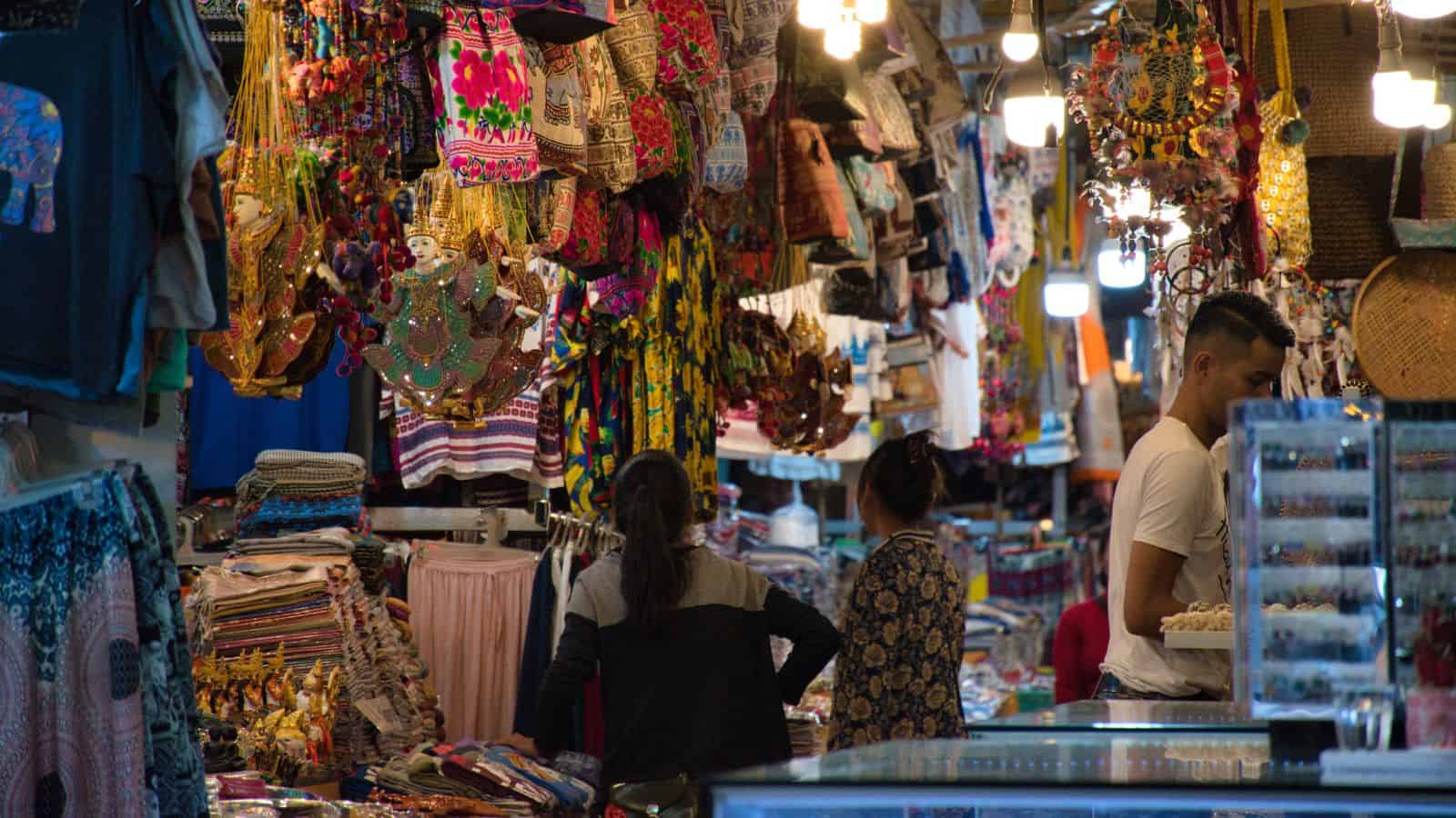Crowded market with colorful textiles, hanging decorations, and people browsing various stalls.