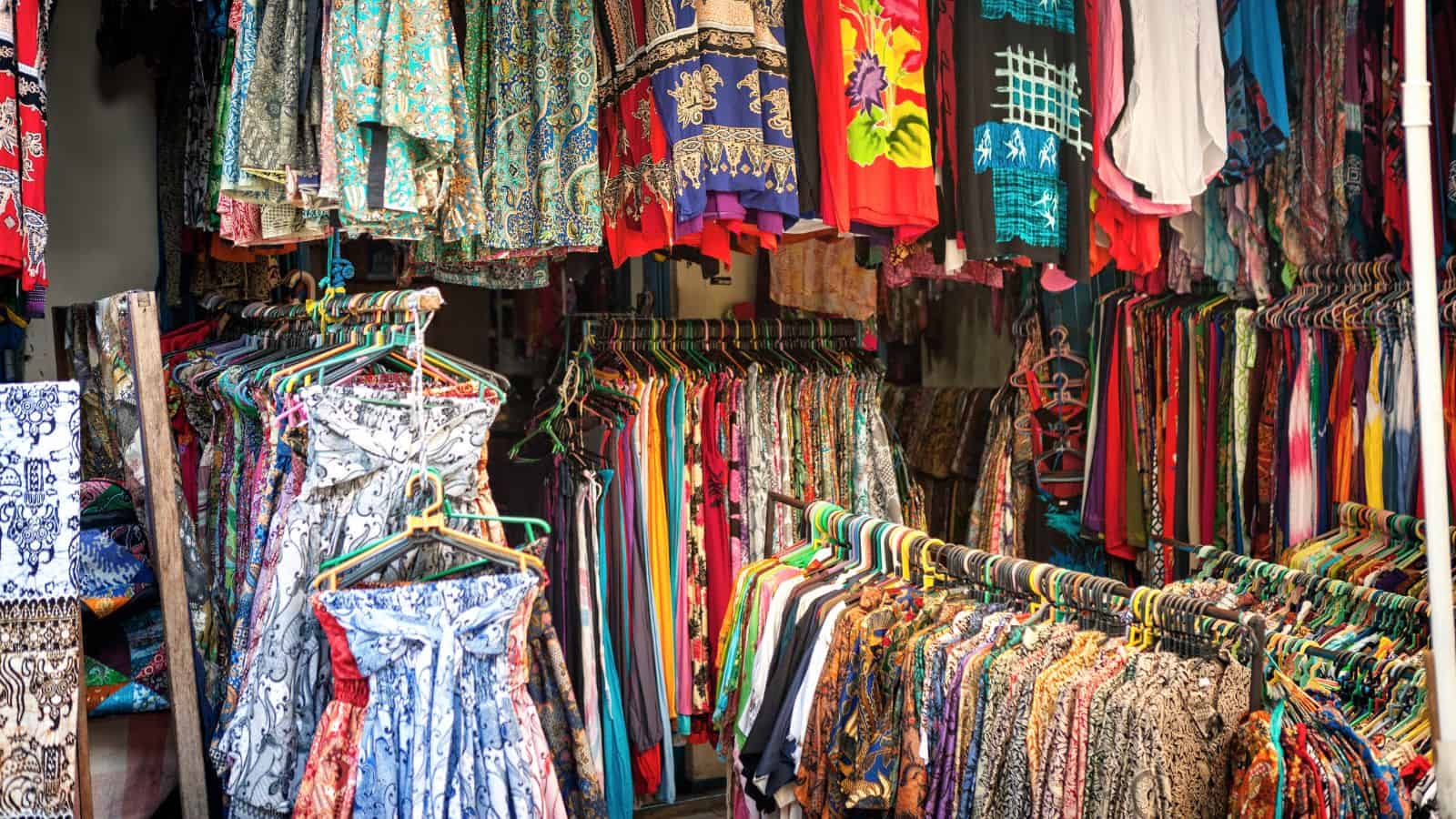 Colorful patterned dresses and shirts hanging on racks in a busy outdoor clothing market.