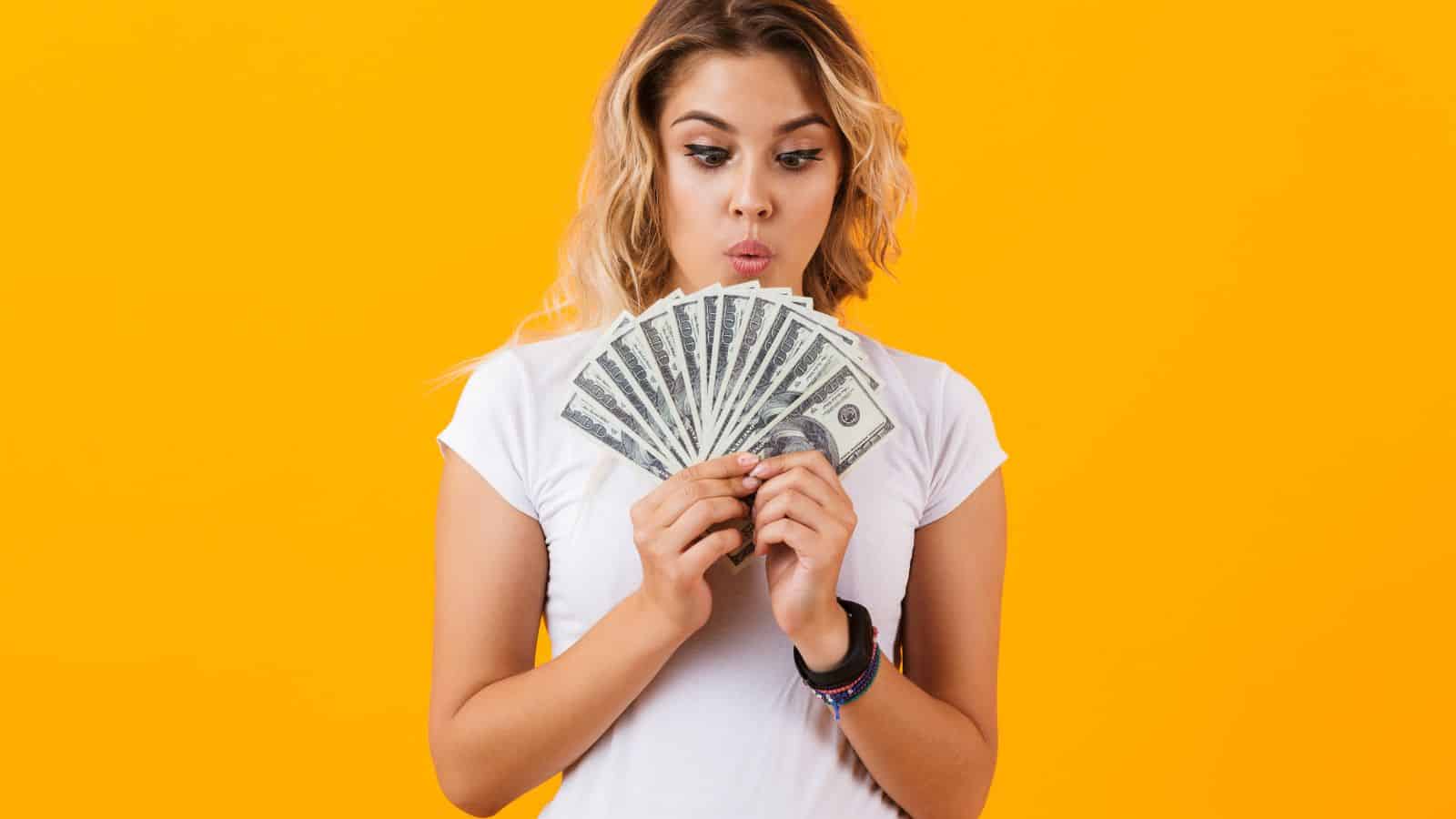 Woman in a white shirt holding a fan of dollar bills, looking surprised, with an orange background.