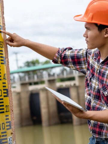 Engineer in orange helmet checks water level gauge at a dam, holding a tablet and pointing at measurement markings.