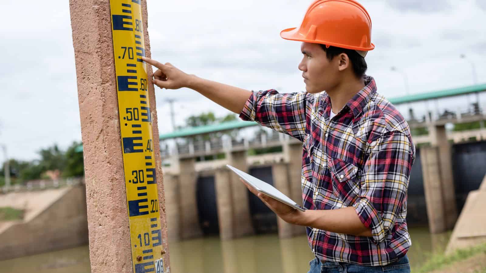 Engineer in orange helmet checks water level gauge at a dam, holding a tablet and pointing at measurement markings.