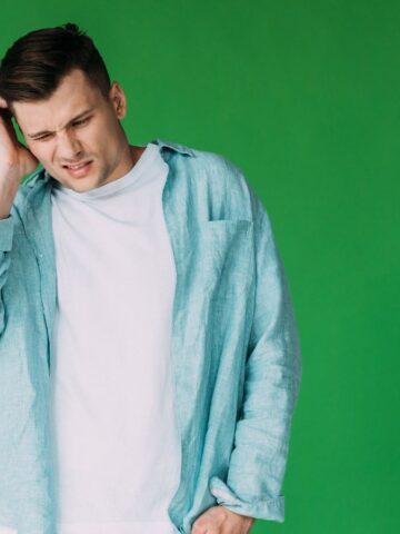 Man in a light blue shirt grimacing and holding his head, standing against a green background.