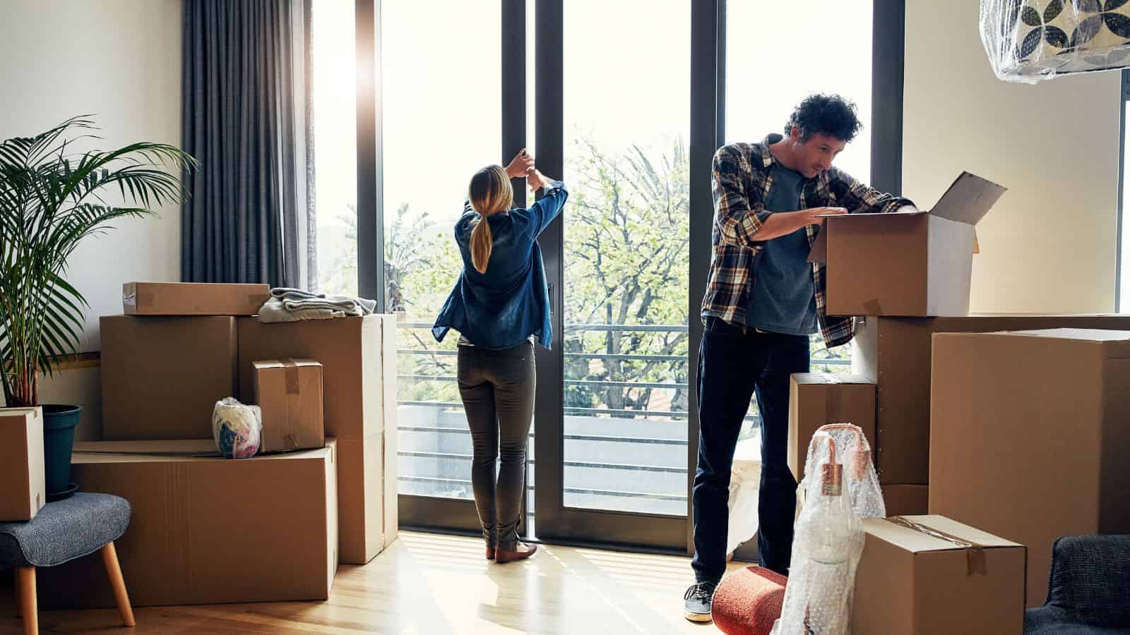 A couple unpacks moving boxes in a bright room with large windows and potted plants.