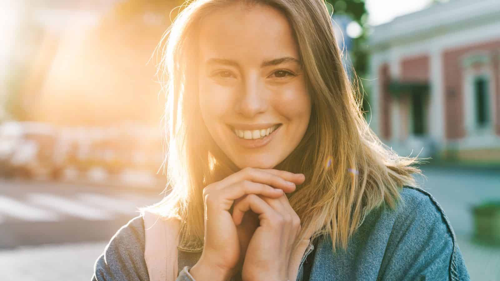 Smiling woman outdoors in sunlight, wearing a denim jacket and holding her hands near her face.