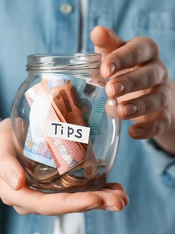 Person holding a glass jar labeled "Tips" filled with cash and coins, wearing a blue shirt.