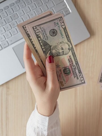Hands holding stacks of U.S. dollar bills near a laptop and a U.S. passport on a wooden desk.