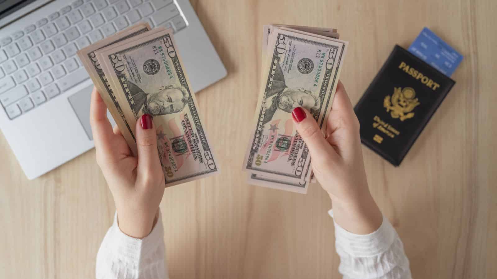 Hands holding stacks of U.S. dollar bills near a laptop and a U.S. passport on a wooden desk.