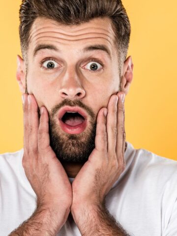Surprised man with beard holding his face, mouth open, against a yellow background—expressing shock like hearing about the water crisis affecting U.S. cities.