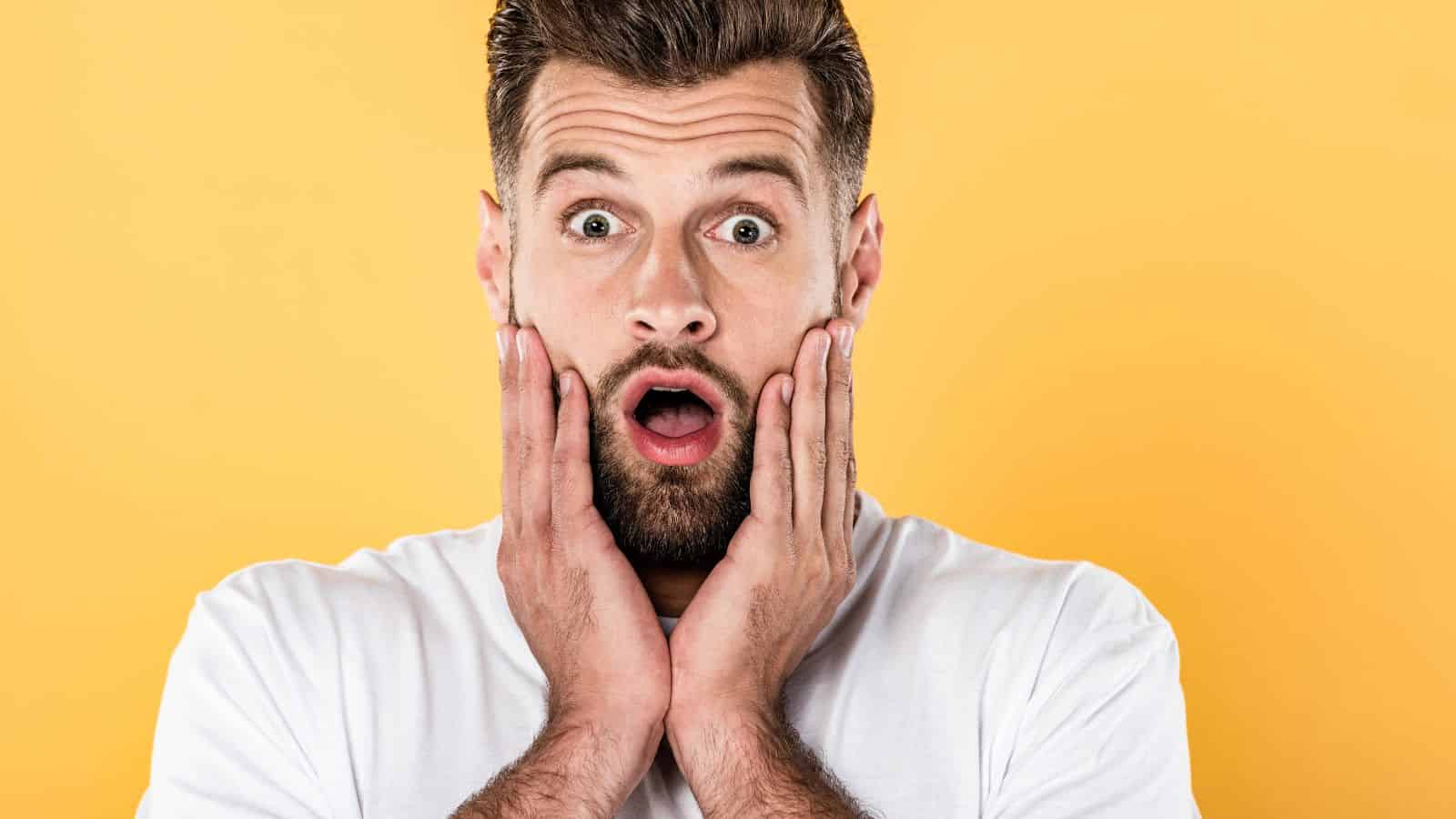 Surprised man with beard holding his face, mouth open, against a yellow background—expressing shock like hearing about the water crisis affecting U.S. cities.