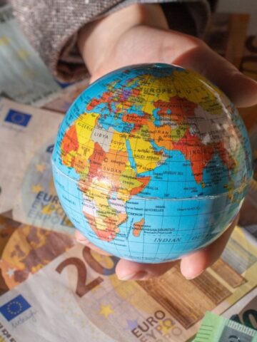 A hand holds a small globe over a scattered pile of euro banknotes of various denominations.