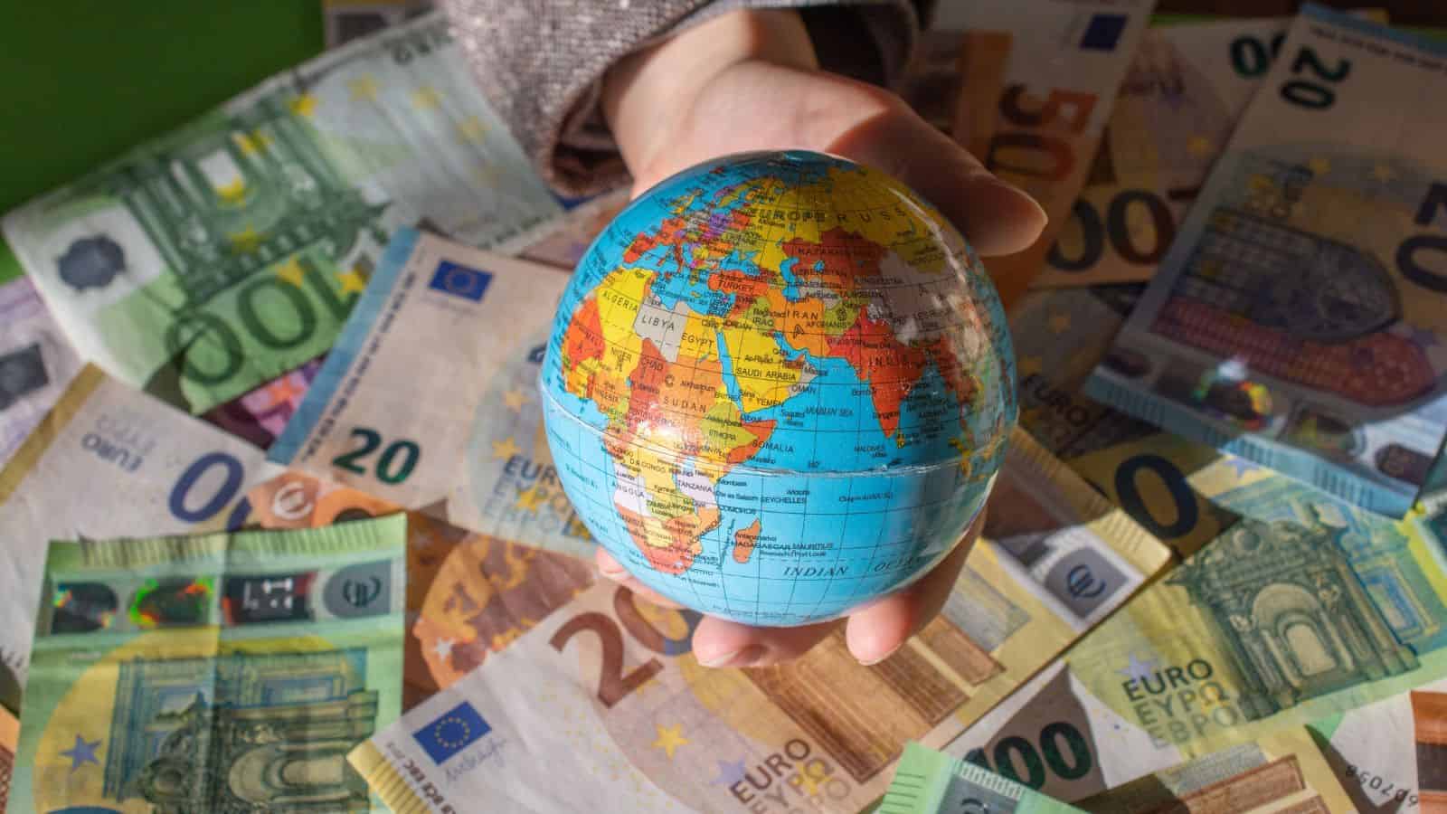 A hand holds a small globe over a scattered pile of euro banknotes of various denominations.