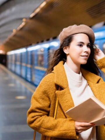 A woman wearing a brown beret and coat holds an open book while standing on a subway platform. A blue subway train is stopped next to her, and the setting appears modern and brightly lit.