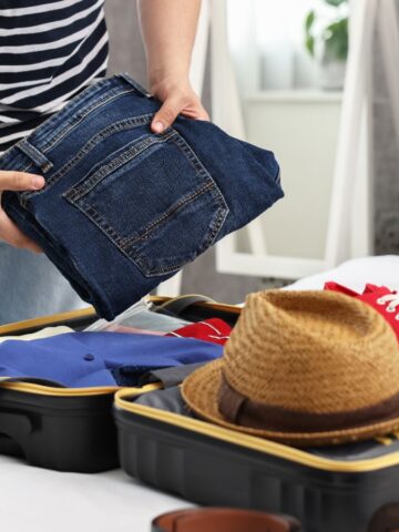 Person packing jeans into an open suitcase on a bed, with a hat, shirt, and red sneakers visible.