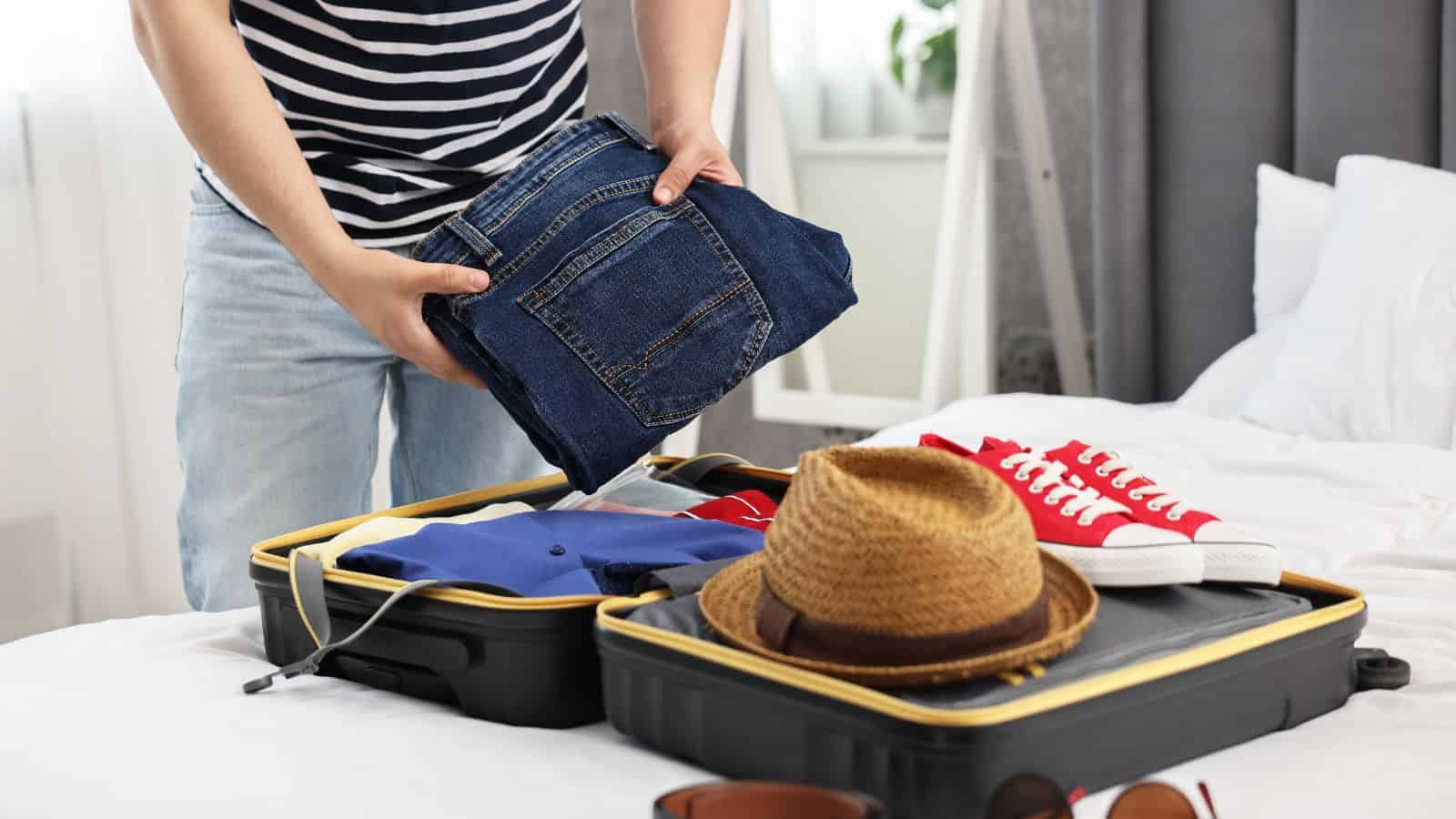 Person packing jeans into an open suitcase on a bed, with a hat, shirt, and red sneakers visible.