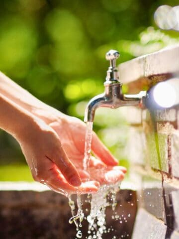 A person holds their hands under running water from an outdoor faucet attached to a brick wall, with green foliage blurred in the background.