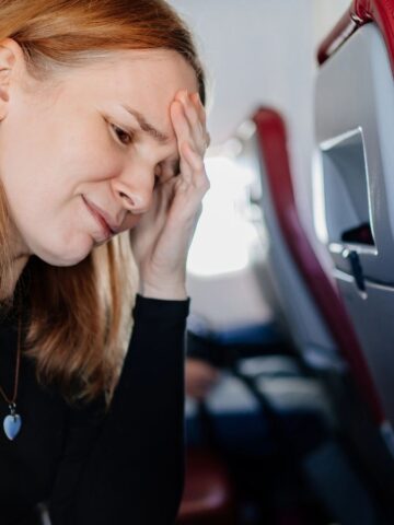 A woman sitting on an airplane holds her forehead, appearing stressed or unwell—a reminder of why it's important to follow travel tips to stay comfortable during your journey.