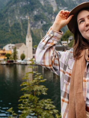 Smiling woman with hat stands by a scenic lakeside village with mountains in the background.