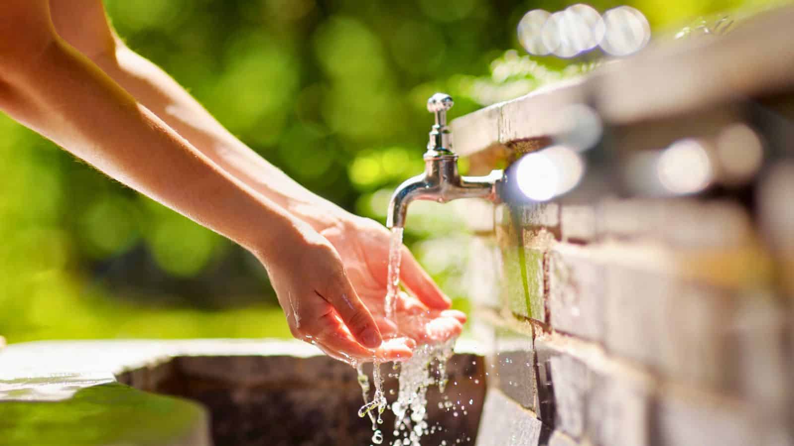 A person holds their hands under running water from an outdoor faucet attached to a brick wall, with green foliage blurred in the background.