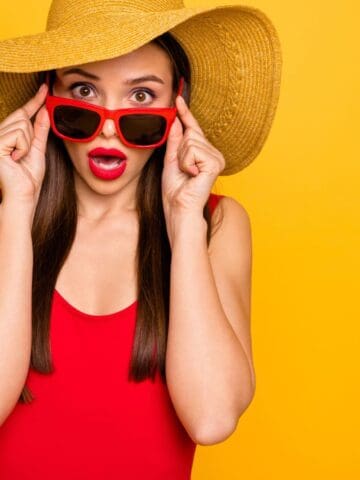 A woman wearing a large straw hat, red sunglasses, and a red top stands against a yellow background, holding her sunglasses with both hands and looking surprised with her mouth open.
