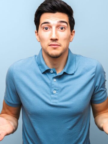 A man in a blue polo shirt shrugs with both hands raised, looking uncertain against a light blue background.