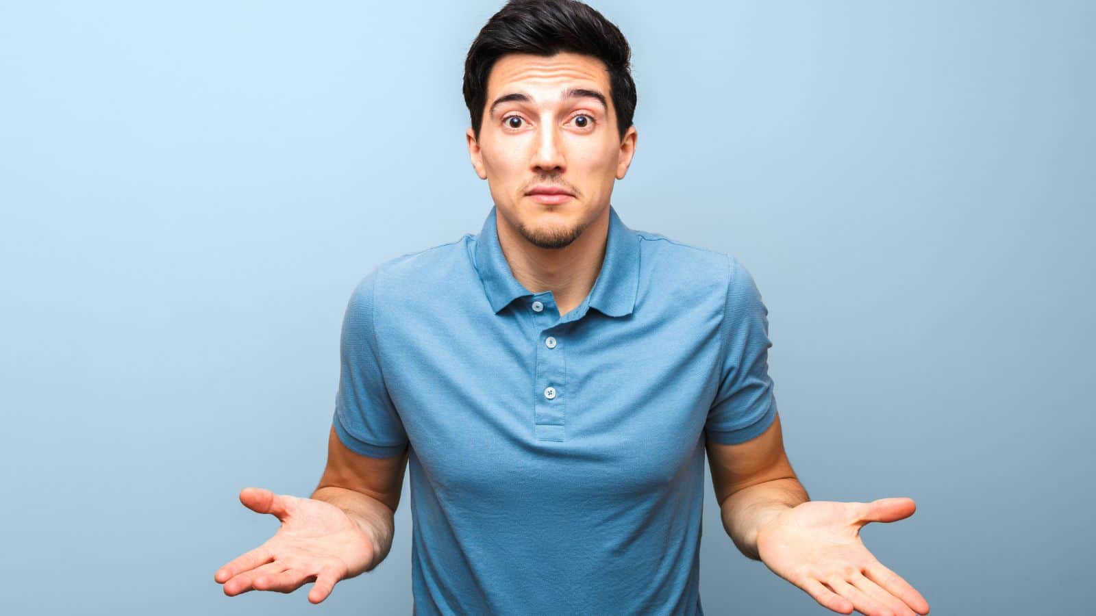 A man in a blue polo shirt shrugs with both hands raised, looking uncertain against a light blue background.