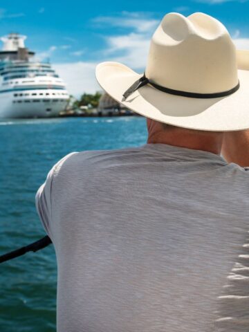 Person in a white cowboy hat looks at cruise ships on the water under a blue sky.
