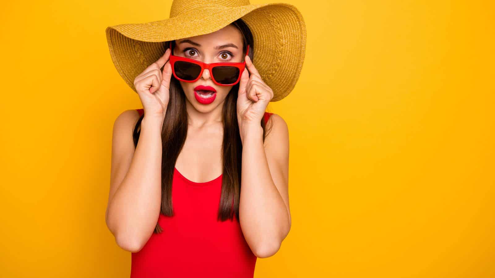 A woman wearing a large straw hat, red sunglasses, and a red top stands against a yellow background, holding her sunglasses with both hands and looking surprised with her mouth open.