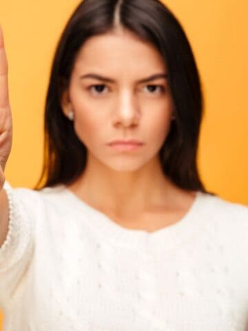 A woman with long dark hair, wearing a white sweater, stands against an orange background. She faces forward with a serious expression, holding up her index finger in front of her.