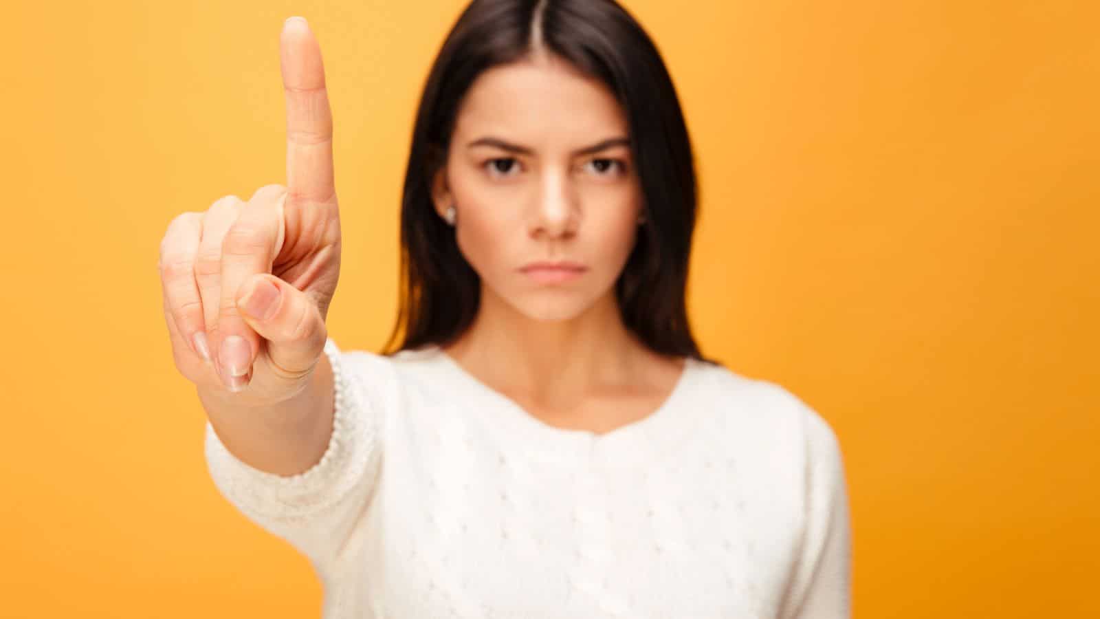 A woman with long dark hair, wearing a white sweater, stands against an orange background. She faces forward with a serious expression, holding up her index finger in front of her.