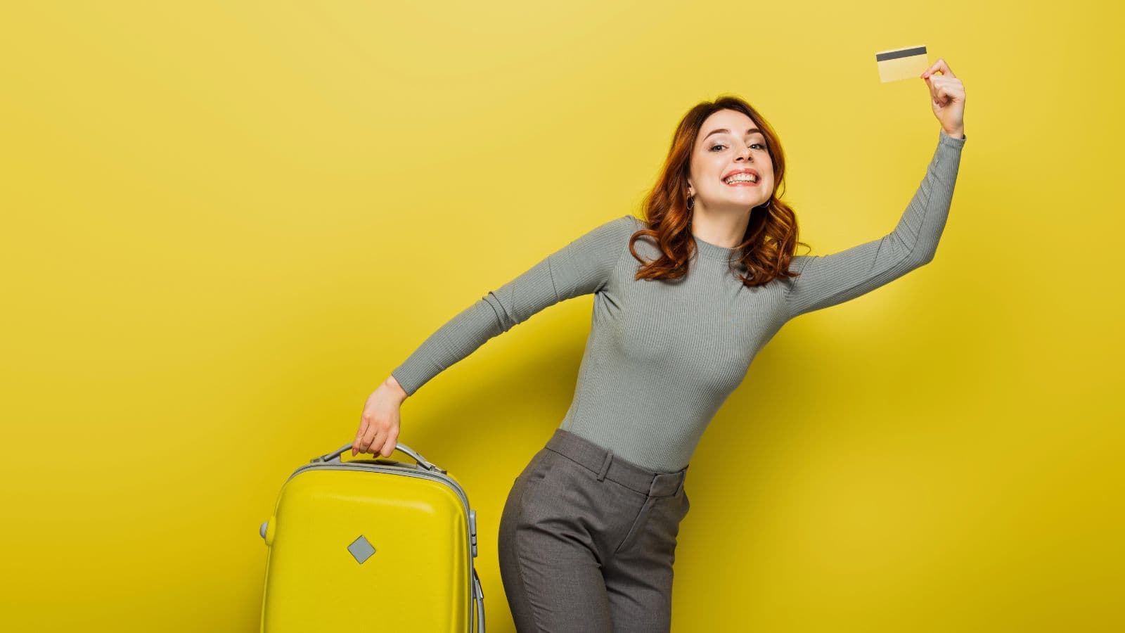 A woman stands against a yellow background, smiling, holding a yellow suitcase with one hand and raising a credit card with the other. She is wearing a grey long-sleeve top and grey pants.
