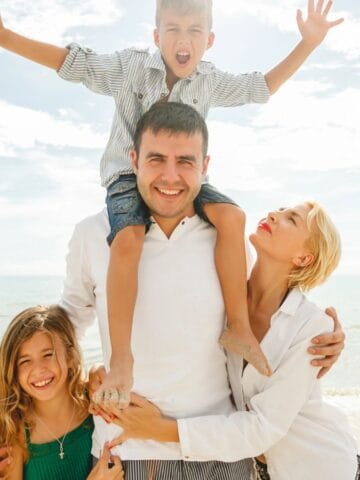 A man stands on a beach with a young boy on his shoulders, a smiling girl to his side, and a woman holding his arm and looking up at him. All appear to be enjoying a sunny day by the sea.