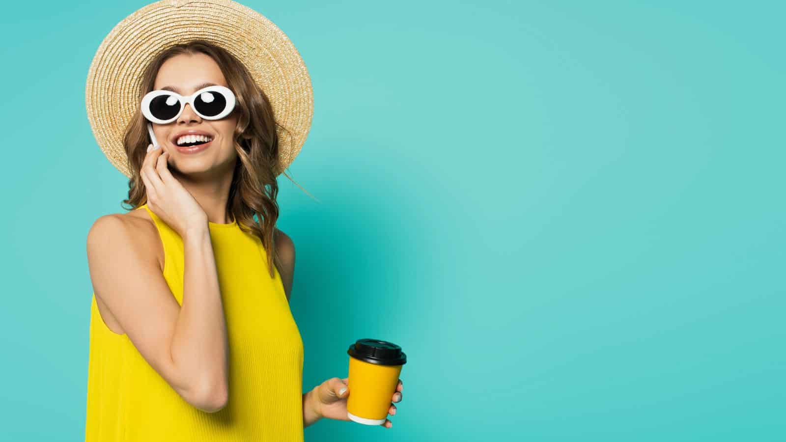 A woman wearing a sunhat, white sunglasses, and a yellow sleeveless top holds a yellow takeaway coffee cup and smiles while touching her face, standing in front of a turquoise background.