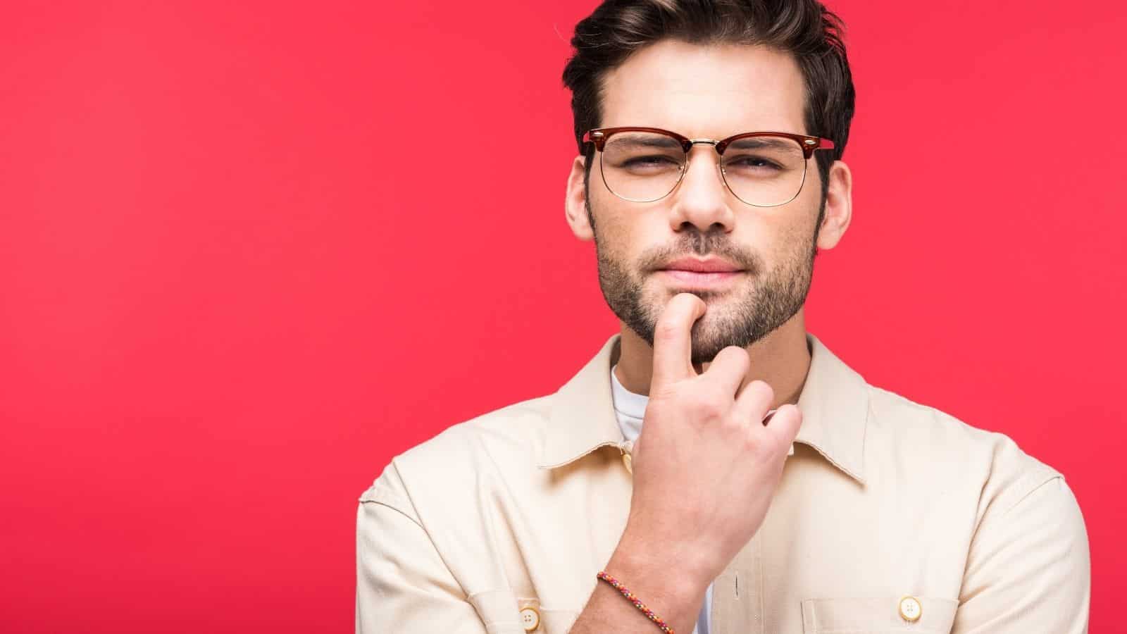 A man with brown hair, glasses, and a trimmed beard stands against a solid red background, touching his chin with a thoughtful expression. He is wearing a light-colored button-up shirt.