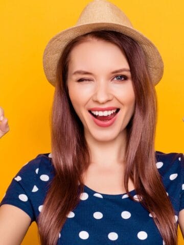 A woman in a straw hat and polka dot shirt smiles, winks, and holds up both hands making "OK" gestures against a solid yellow background.