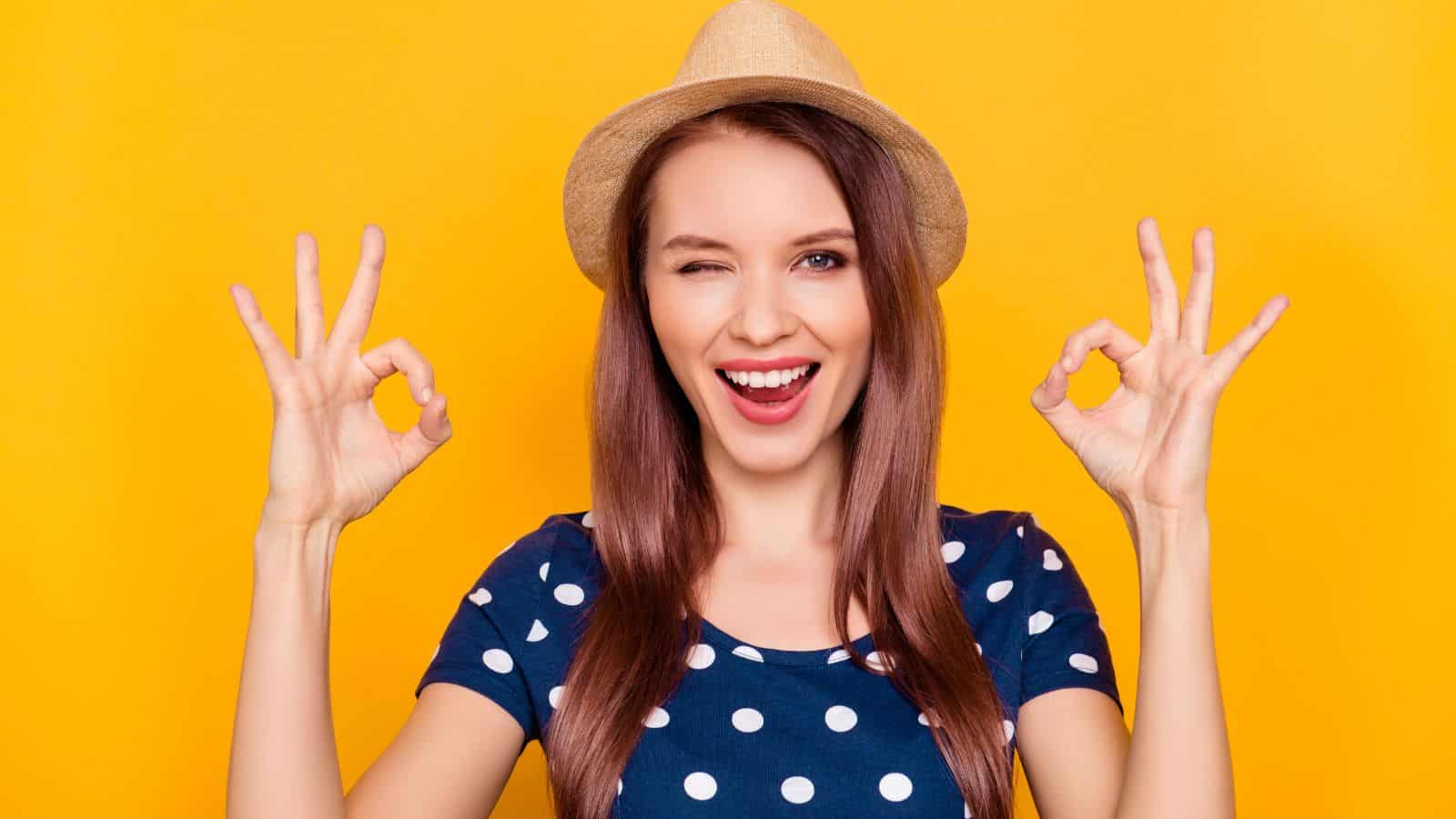A woman in a straw hat and polka dot shirt smiles, winks, and holds up both hands making "OK" gestures against a solid yellow background.