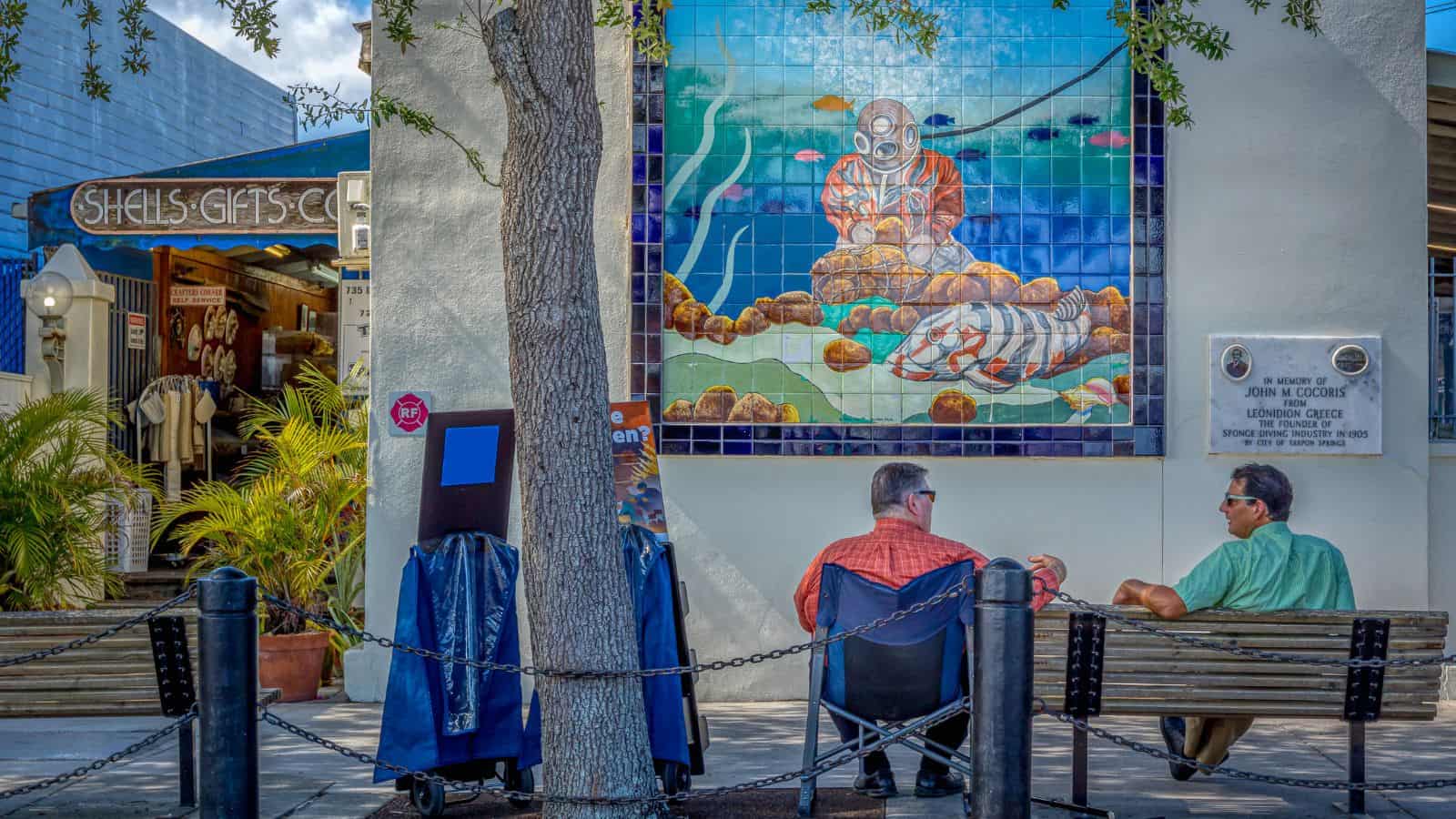 Two men sit on benches under a tree, chatting in an American town. Behind them, a large tile mural of a deep-sea diver with lobsters adds European style flair, while "Shea's Gifts" welcomes travelers from the nearby shop entrance.