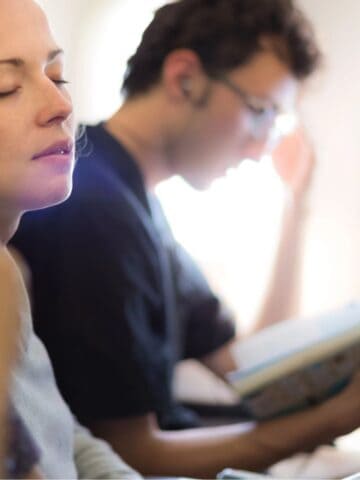 Three people sit in an airplane row. The person closest to the camera rests their chin on their hand. The middle person has their eyes closed and appears relaxed. The farthest person reads a book and wears wired earphones.