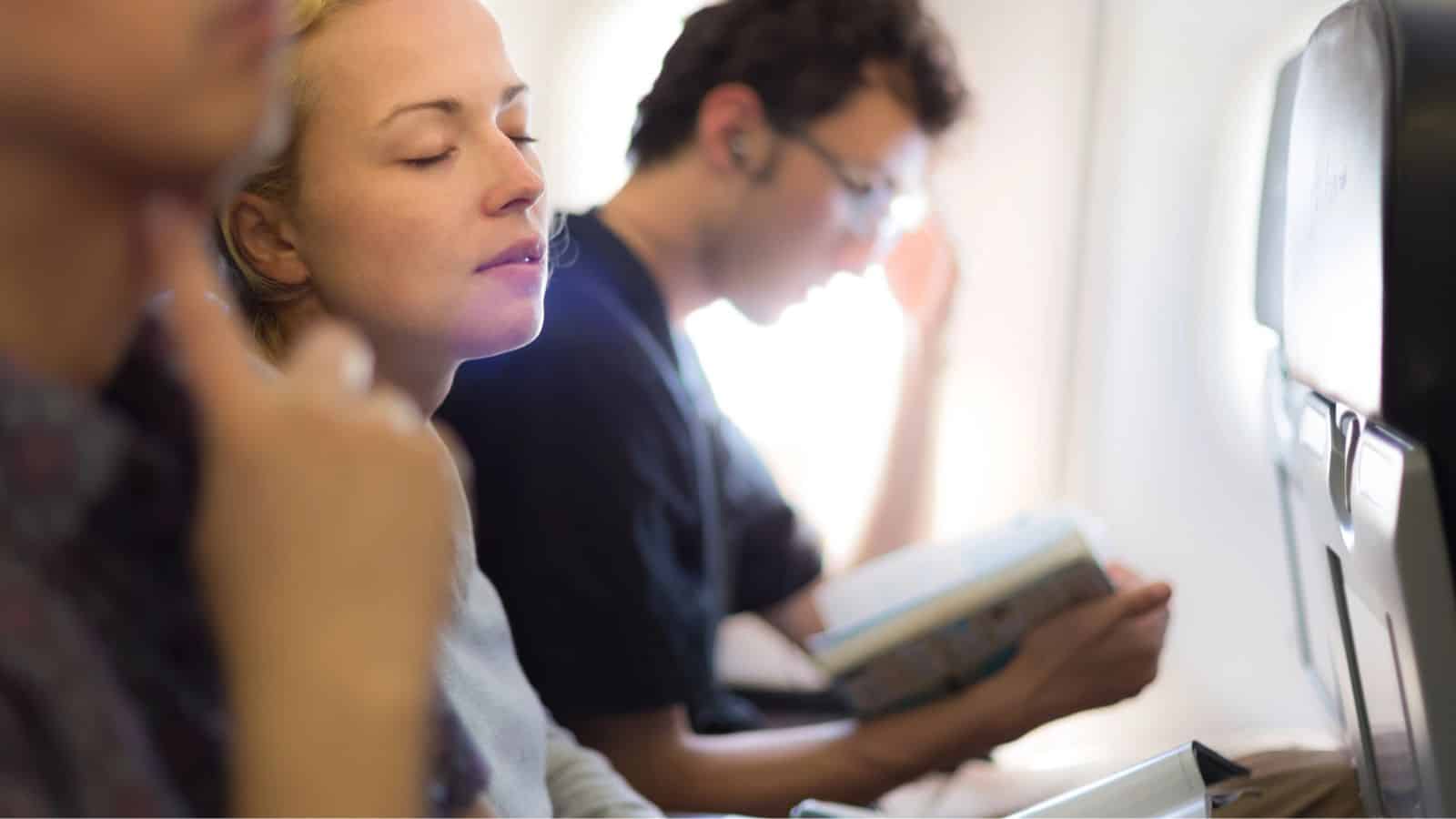 Three people sit in an airplane row. The person closest to the camera rests their chin on their hand. The middle person has their eyes closed and appears relaxed. The farthest person reads a book and wears wired earphones.
