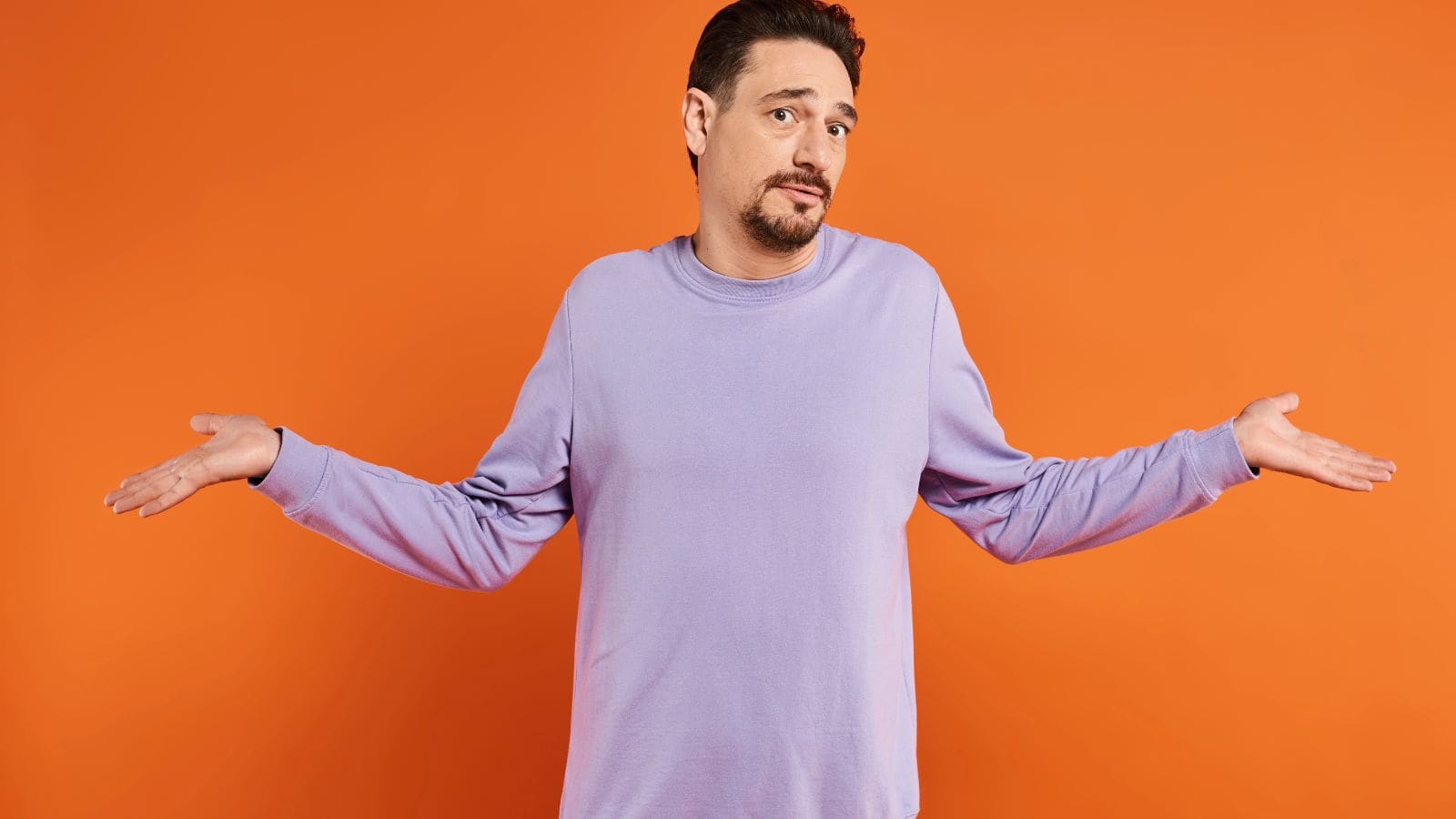A man wearing a light purple long-sleeve shirt stands in front of an orange background with his arms outstretched and palms up, appearing to shrug or indicate uncertainty.