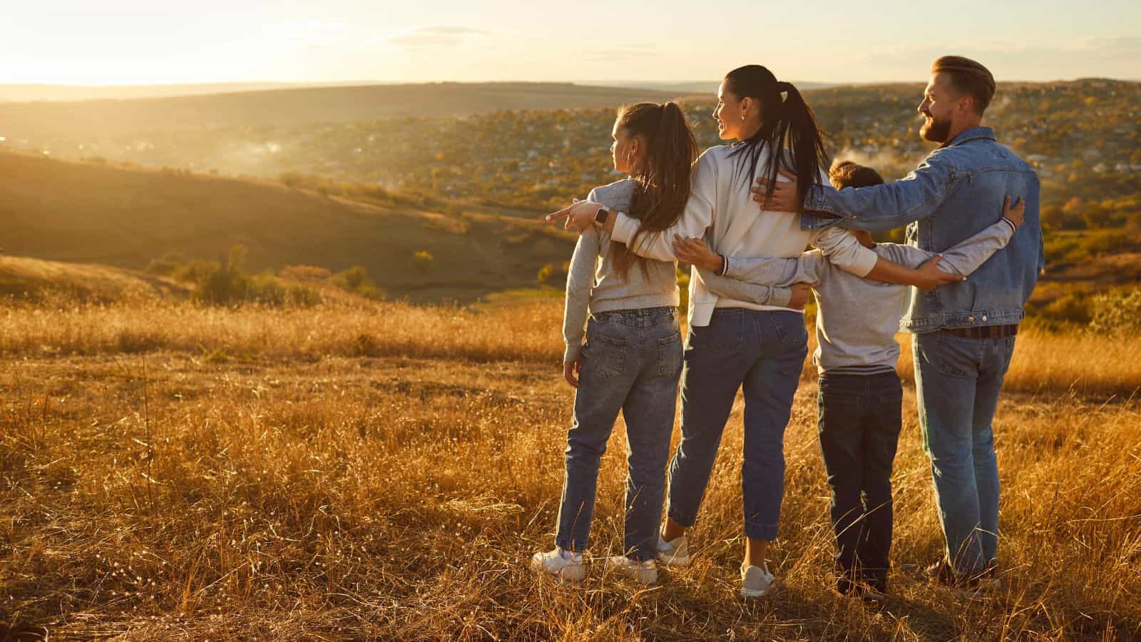 Four people, two adults and two children, stand close together in a grassy field, facing away from the camera and looking out over a hilly landscape at sunset or sunrise.