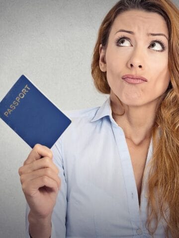 A woman with long hair in a light blue shirt holds a blue passport in one hand and a red passport in the other, looking upward with a puzzled expression against a plain, gray background.