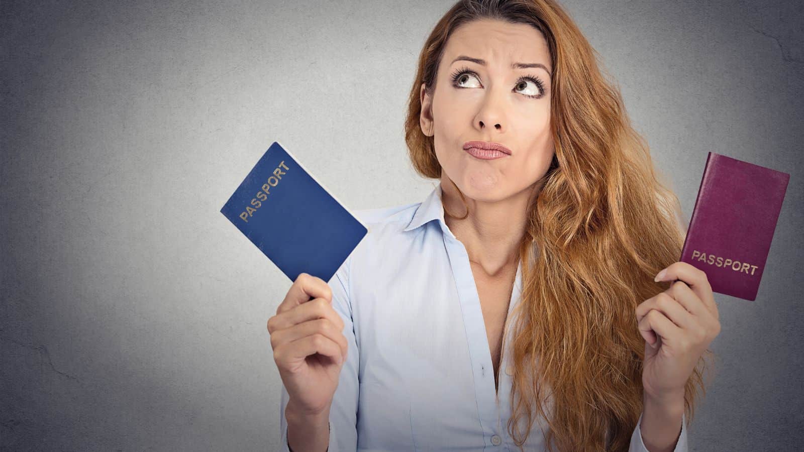 A woman with long hair in a light blue shirt holds a blue passport in one hand and a red passport in the other, looking upward with a puzzled expression against a plain, gray background.