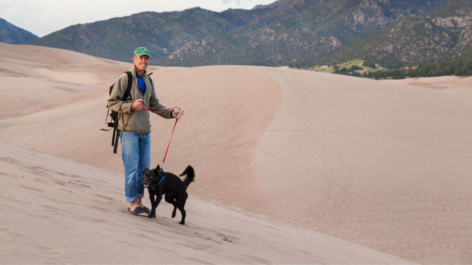 A person wearing a green cap, jacket, and rolled-up jeans walks a black dog on a red leash across sand dunes with mountains and greenery visible in the background.