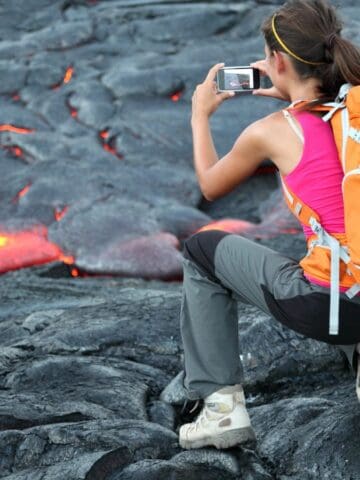 Two hikers with backpacks crouch on hardened black lava, with one taking a photo of glowing orange molten lava nearby. Both wear outdoor clothing and appear focused on the lava flow.