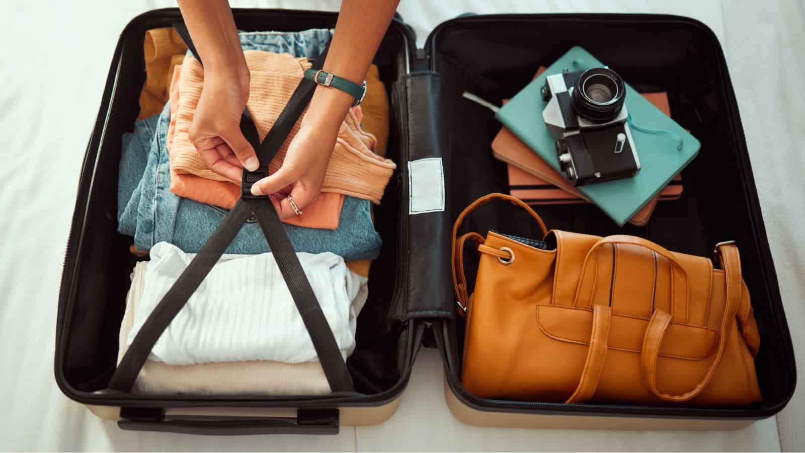 A person secures the straps inside an open suitcase filled with folded clothes, a camera, notebooks, and a brown handbag on a bed—showcasing packing tips to make the most of your carry-on space.