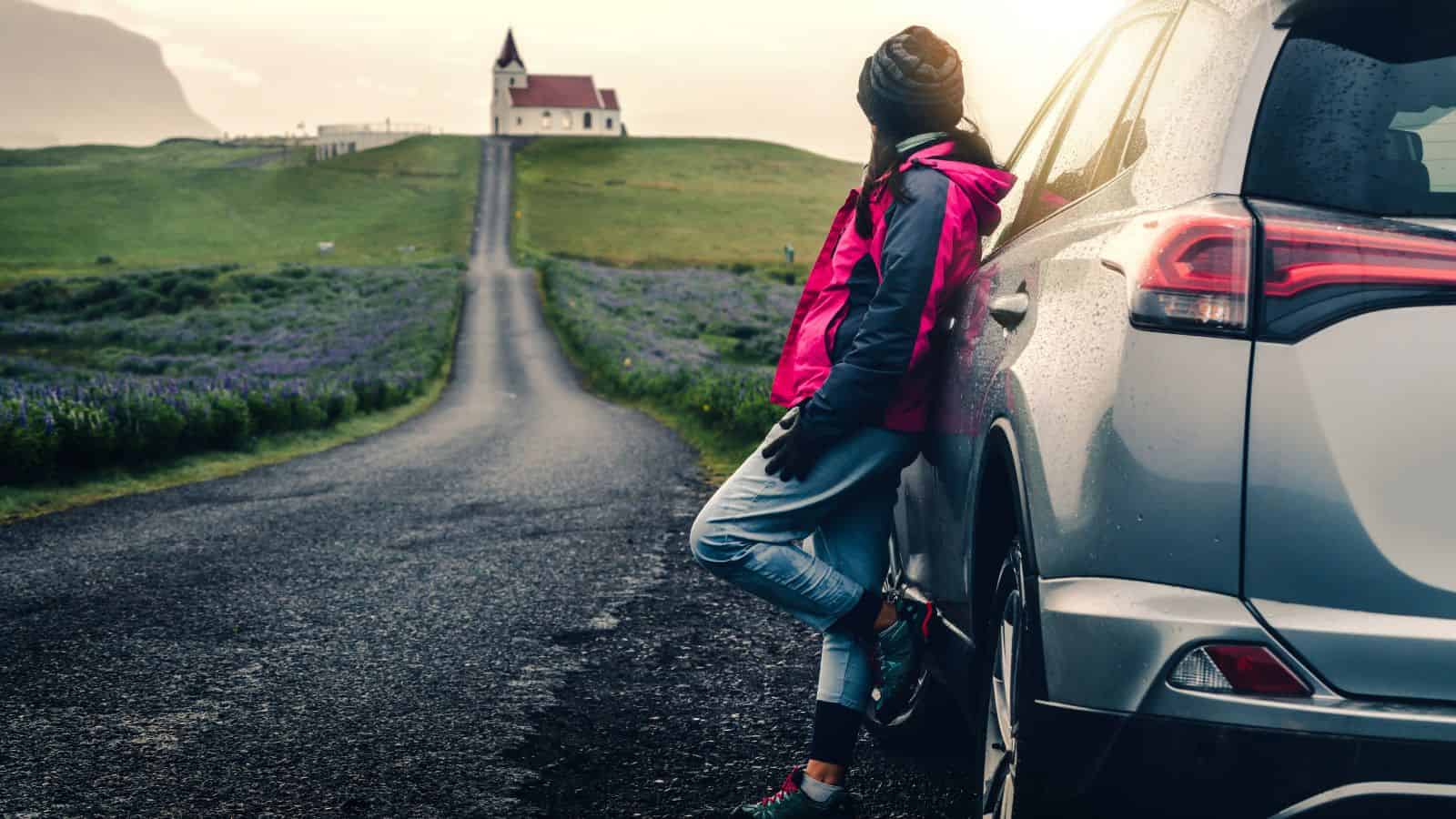 Person in a pink jacket leans on a car, gazing at a distant church at the end of a country road.